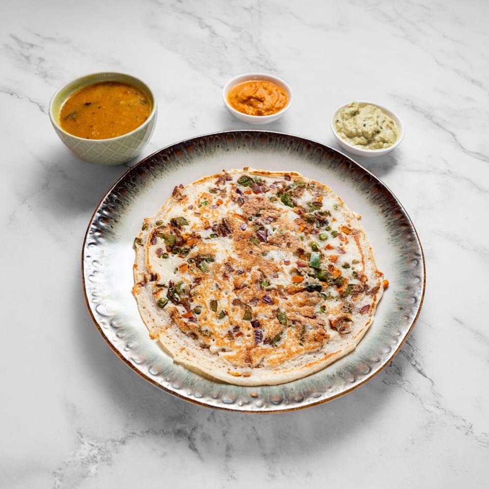 Masala Symphony's Indian uttapam with vegetables, served with sambar and two chutneys on a marble surface.