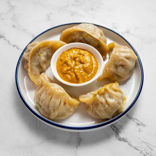 A plate of steamed momos with a small bowl of orange dipping sauce on a marble surface.