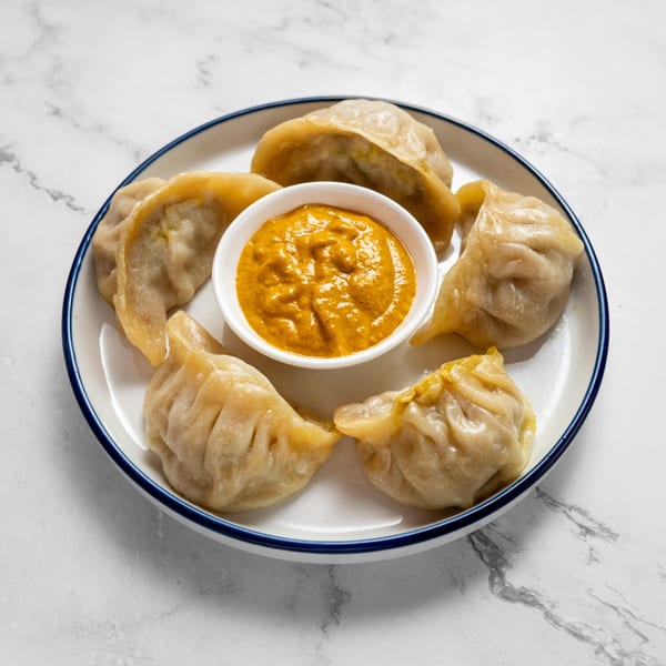 A white plate with blue rim holds six steamed momos and a small bowl of orange dipping sauce.