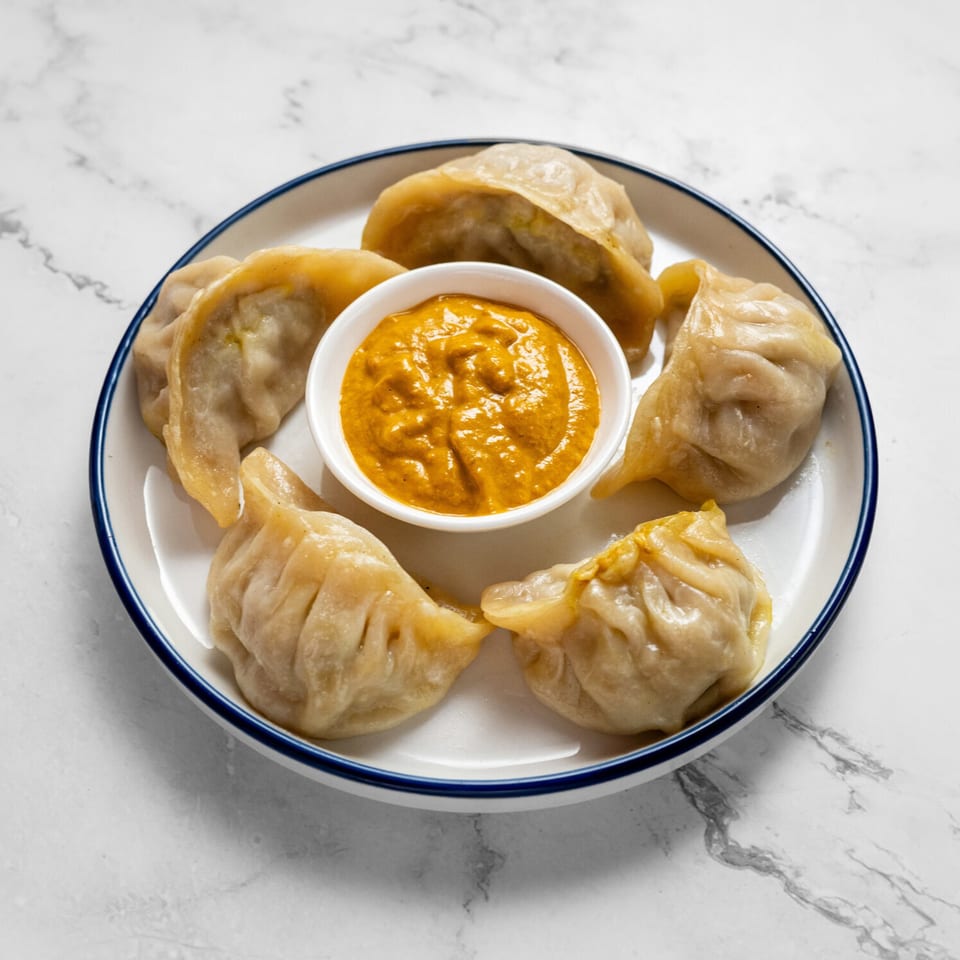 A white plate with blue rim holds six steamed momos and a small bowl of orange dipping sauce.