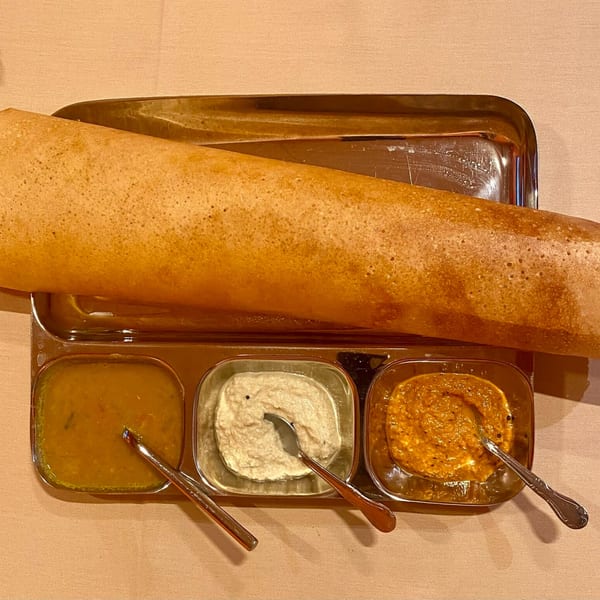 A golden-brown dosa on a steel tray with three small bowls of Indian chutneys and sambar.