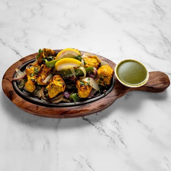 Indian chicken tikka sizzler with onions, peppers, lemon, and green chutney on a marble surface.