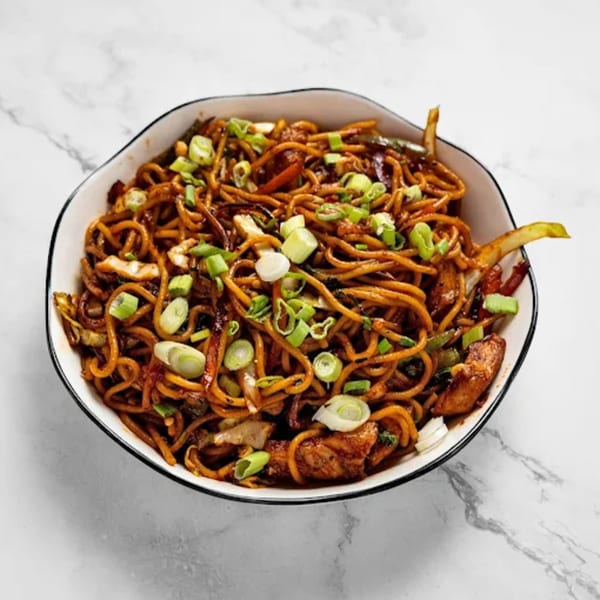 A bowl of Masala Symphony's chicken noodles, garnished with chopped green onions, on a marble surface.