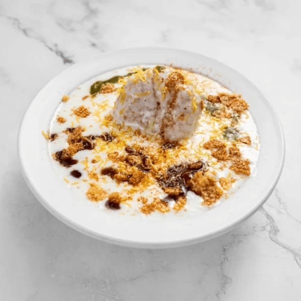 A white bowl of Dahi Bhalla, topped with yogurt, chutneys, and crunchy sev on a marble surface.